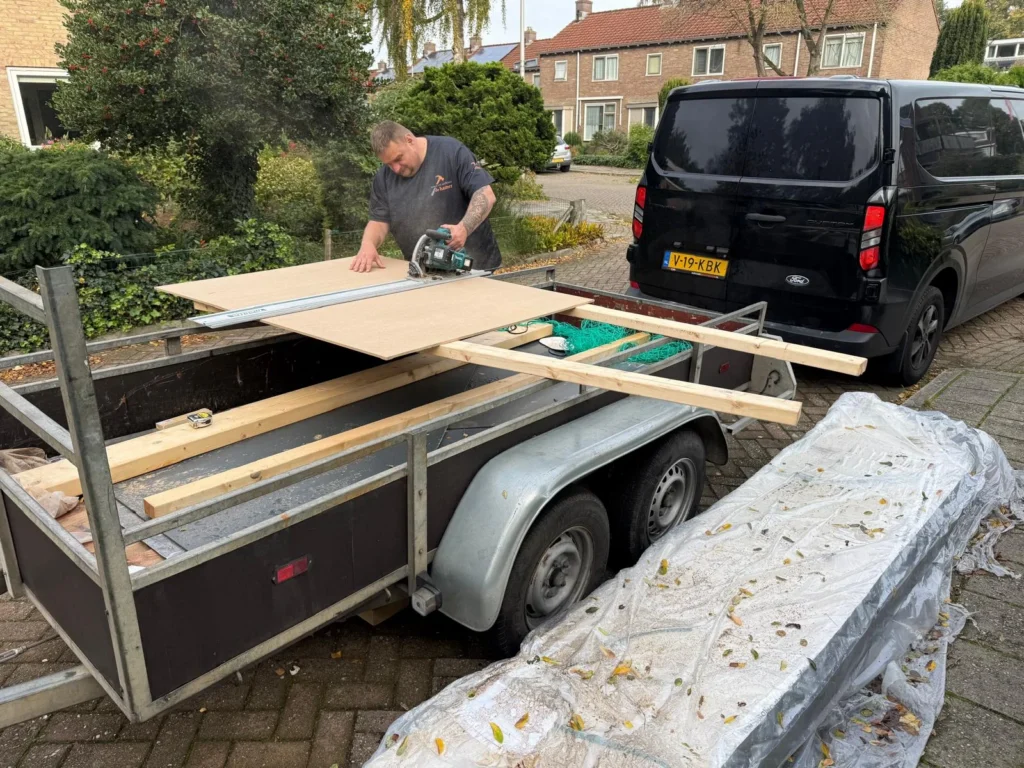 Vakman zaagt plaatmateriaal op een aanhanger naast een zwarte bestelbus in Apeldoorn