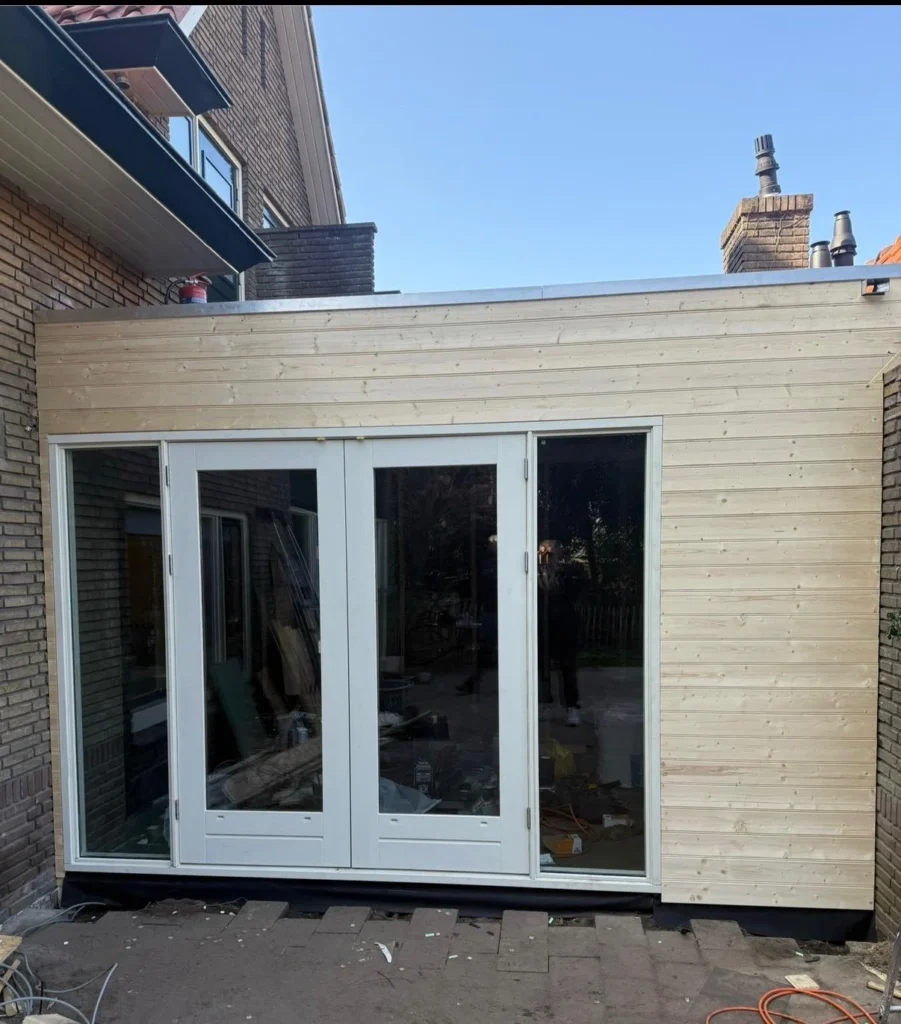 Nieuwe uitbouw met houten gevelbekleding, plat dak en witte openslaande deuren in Apeldoorn